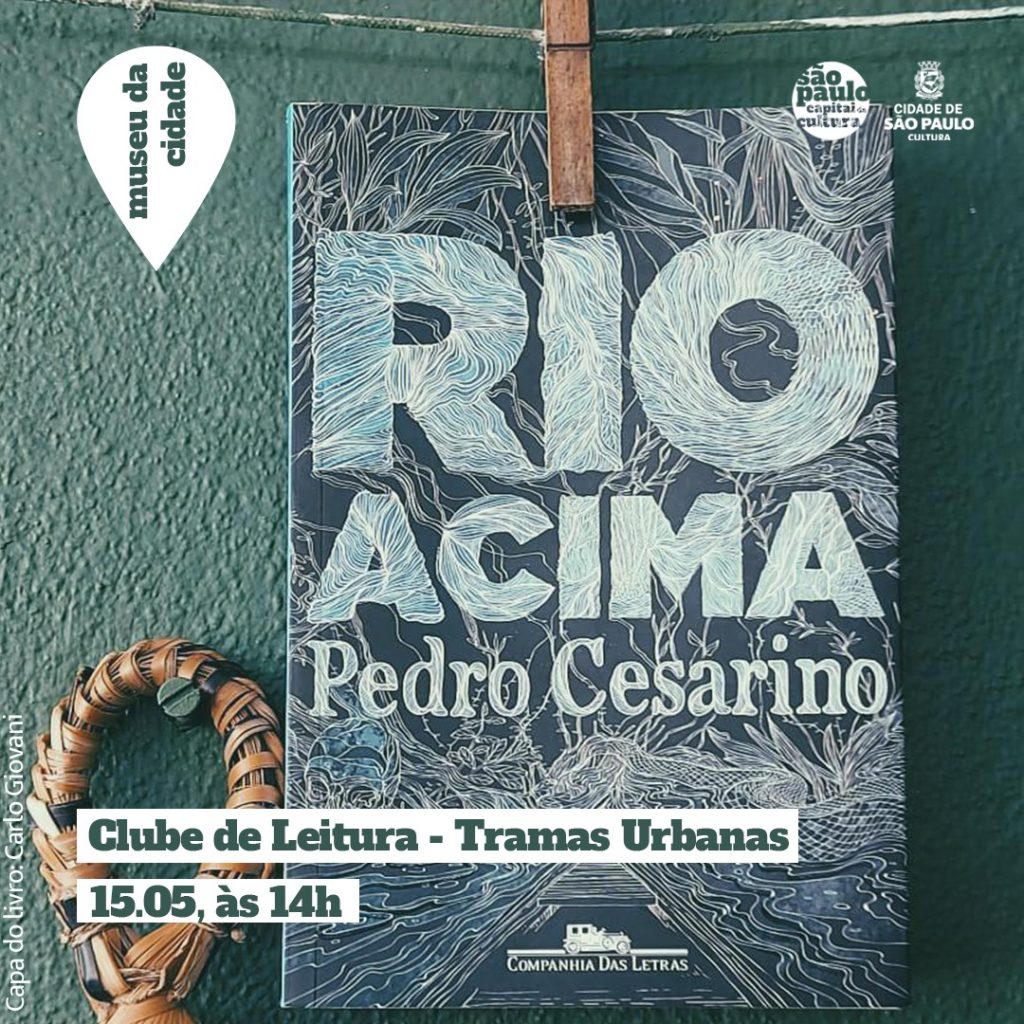 Foto tirada em direção a parede. Pendurado no varal, por um pregador de madeira, o livro “Rio Acima”. Ao lado, um objeto de palha pendurado em um parafuso. Ao fundo, uma parede verde. Na foto está escrito “Clube de Leitura | Tramas Urbanas 15.05, às 14h". “ Capa do livro: Carlo Giovani”