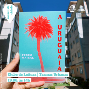 Entre prédios em ambos os lados, segurado por uma mão, vemos o livro “A Uruguaia” com um coqueiro em sua capa e o título na vertical ao lado direito. Acima um céu claro compõe a fotografia. Nela, está escrito “Clube de Leitura | Tramas Urbanas 19.06, às 14h. Capa: Julia Masagão e Tali Kimelman”