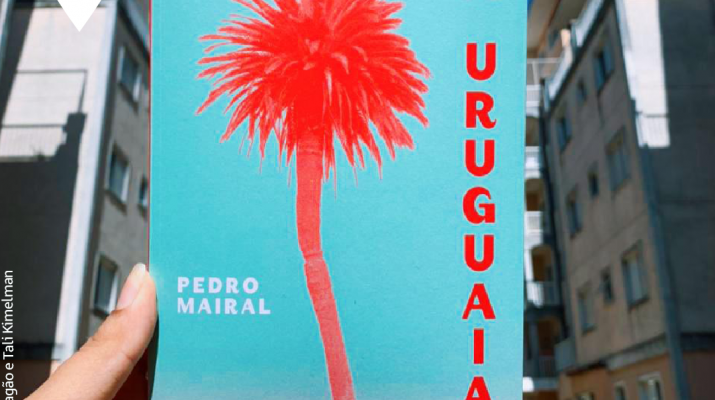 Entre prédios em ambos os lados, segurado por uma mão, vemos o livro “A Uruguaia” com um coqueiro em sua capa e o título na vertical ao lado direito. Acima um céu claro compõe a fotografia. Nela, está escrito “Clube de Leitura | Tramas Urbanas 19.06, às 14h. Capa: Julia Masagão e Tali Kimelman”